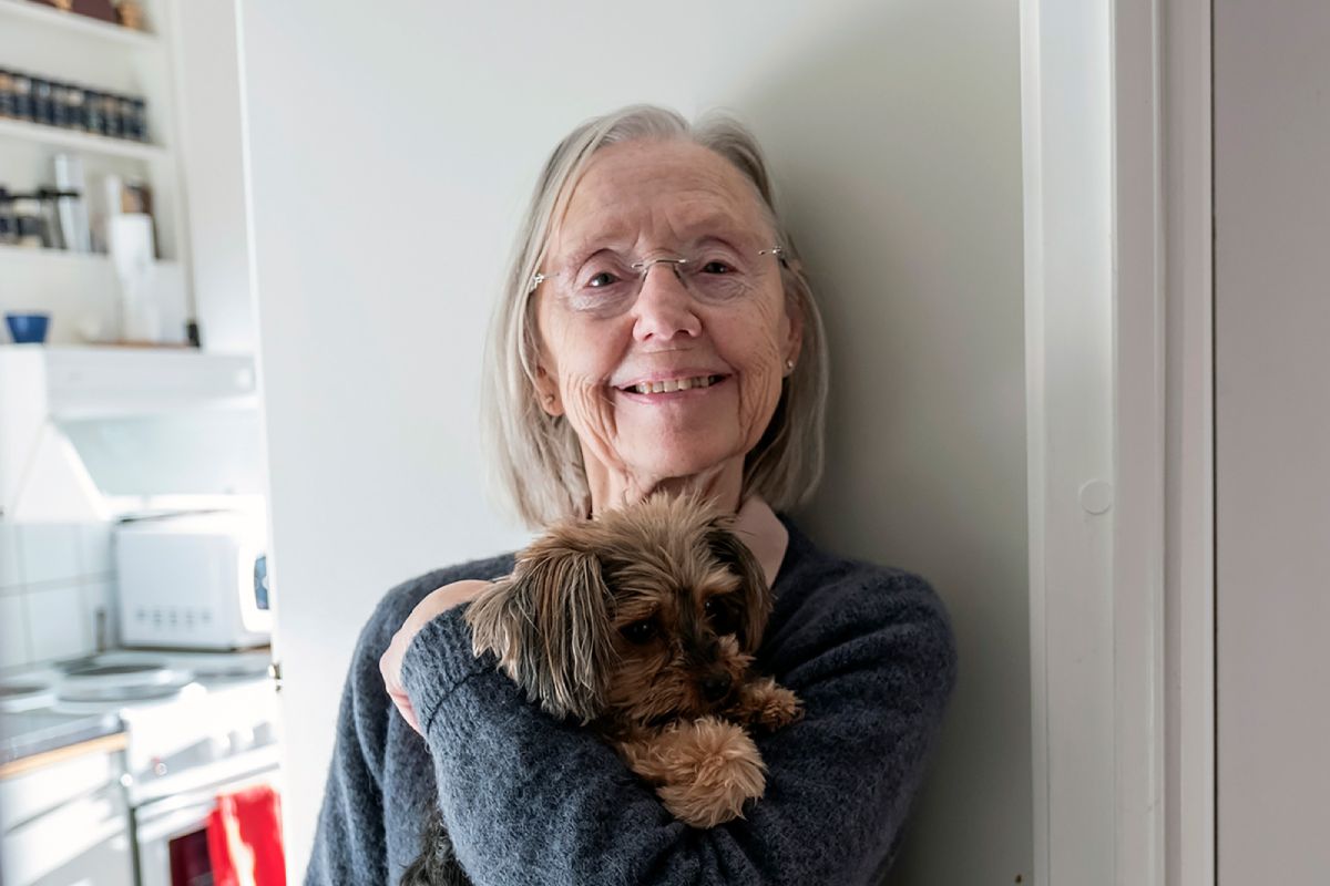 En glad äldre kvinna håller en hund i sin famn och tittar in i kameran.