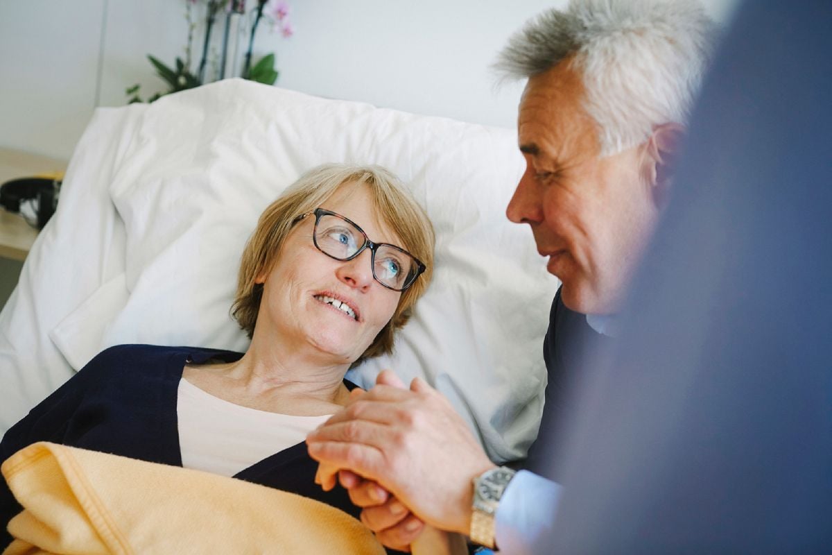 En kvinna ligger i en sjukhussäng och pratar med en man, som håller henne i handen.