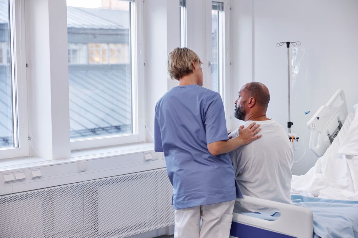 En man sitter på en sjukhussäng. En sjuksköterska lägger sin hand på hans axel.